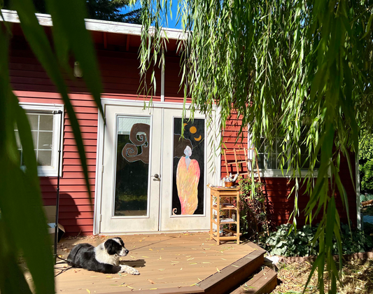 Red Art studio with a painting of a woman on the door.  A black and white dog is laying on the deck and willow branches are hanging around the frame of the photo