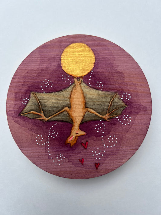 Round painting of a bat hanging upside down from a gold full moon.  The background is purple with white, swirly dots of white and three small red hearts.